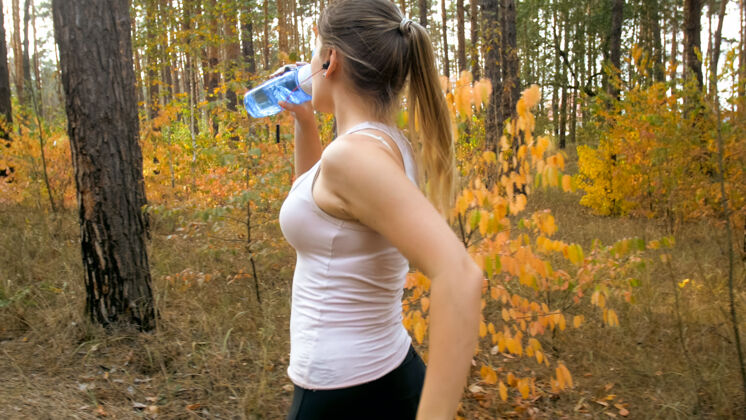 自然年轻的金发女人在森林里奔跑 喝着瓶子里的水的画像马拉松乡村路径