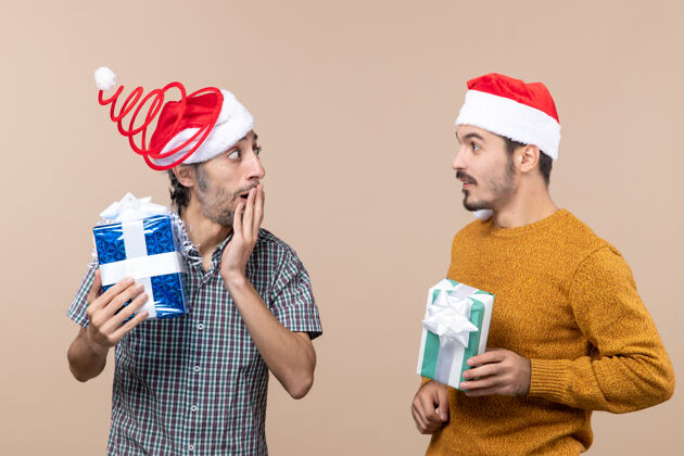 持有前视图两个困惑的家伙戴着圣诞帽拿着圣诞礼物分享秘密米色孤立的背景帽子帽子成人
