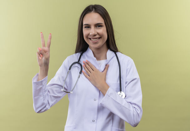 听诊器带着微笑的年轻医生女孩穿着听诊器医用长袍展示和平的姿态把她的手放在绿色背景的心脏上年轻手势医生