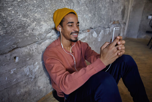 裤子正面正面的深色胡须 穿着粉色毛衣 蓝色裤子 裤子和芥末帽的家伙坐在木地板上 手里拿着智能手机 戴着耳机听音乐视频非洲年轻