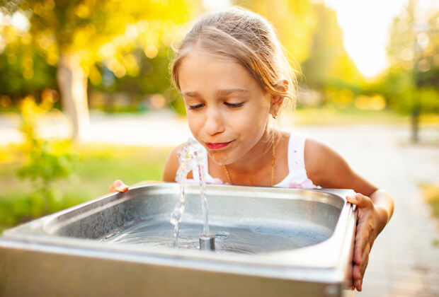 阳光在期待已久的假期里 一个快乐的好女孩在一个夏日阳光明媚的公园里 从一个小喷泉里喝着清凉的淡水水龙头排水关系