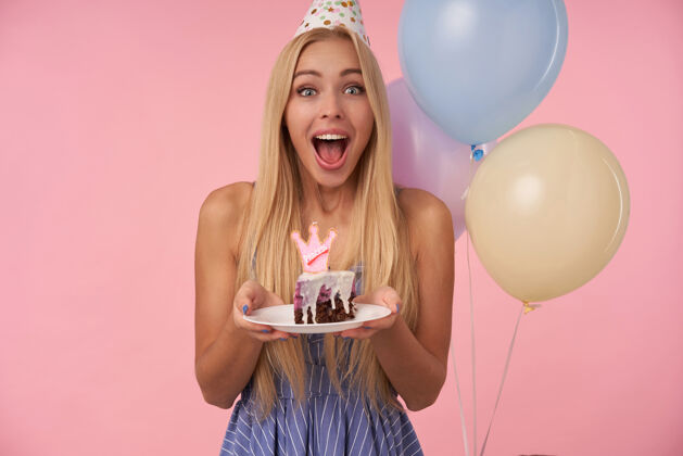 头发快乐的年轻漂亮女人 长着金色的头发 穿着蓝色的夏装 戴着圆锥形的帽子 庆祝生日 手里拿着一块蛋糕 在粉色的背景下露出灿烂的笑容发型迷人节日