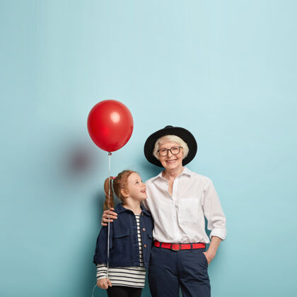 拥抱家庭庆典概念可爱的红发女孩用母亲节祝贺成熟的老奶奶 捧着红色气球 拥抱在一起 隔着空白的蓝色墙壁老人奶奶眼镜