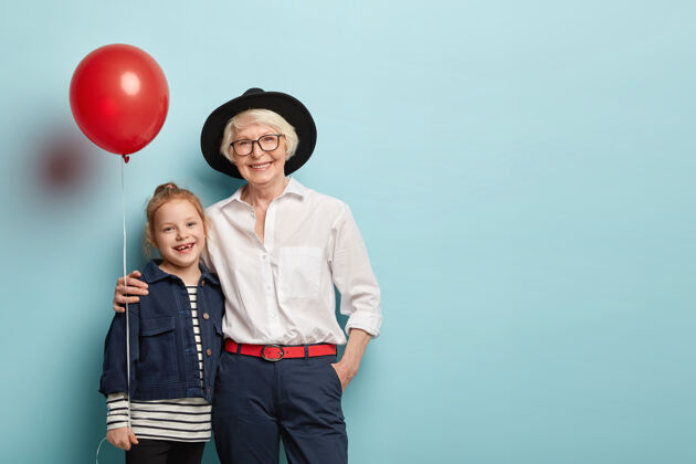 拥抱孙女和奶奶的家庭肖像拥抱和庆祝节日 手持气球 穿着喜庆的衣服 表达积极的情绪隔离在蓝色的墙壁上一代人和节日的概念高兴女人皱纹