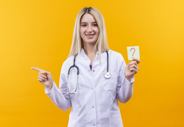 女孩微笑的医生年轻的女孩穿着医用长袍戴着听诊器和牙套拿着纸问号指向隔离的黄色背景边问题举行点
