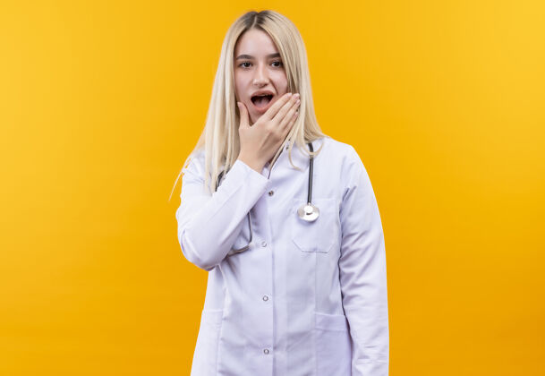 听诊器悲伤的医生穿着医用长袍戴着听诊器的年轻女孩把手放在孤立的黄色背景上疼痛的牙齿上悲伤牙齿疼痛