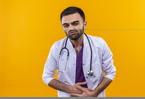 疼痛生病的年轻男医生穿着听诊器医用长袍 在孤立的黄色背景上抓住了疼痛的胃胃长袍听诊器