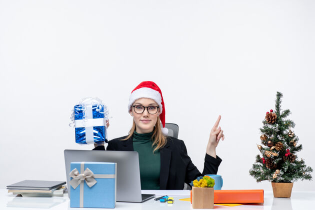 礼物圣诞气氛与戴着圣诞老人帽子和眼镜的年轻女子坐在一张桌子旁 拿着她的礼物 指着上面的白色背景拿着坐着女人