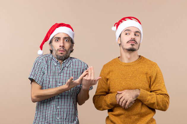 帽子两个戴着圣诞帽的家伙在米色孤立的背景下冒犯了我情侣圣诞老人男