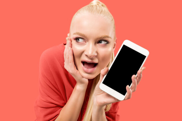 软件一个惊讶的 微笑的 快乐的 惊讶的女孩的肖像 显示在珊瑚背景下的空白屏幕手机电话金发网络
