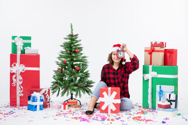 前面前视图的年轻女子坐在圣诞礼物周围拿着白色墙上的时钟庆祝坐着圣诞快乐
