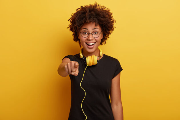 耳机可爱的积极的黑皮肤女孩穿着黑色t恤 圆眼镜 有黄色的耳机连接到一些小玩意 是真正的梅洛曼播放列表音频青少年