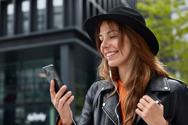 满足横向拍摄的高兴的女模特在时尚的黑色帽子和皮夹克 集中在智能手机设备科技手机积极