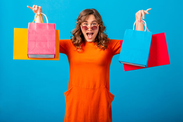 年轻图片中的兴奋迷人的微笑时尚女性购物狂穿着橙色的新潮连衣裙拿着购物袋在蓝色的工作室背景隔离包购买太阳镜