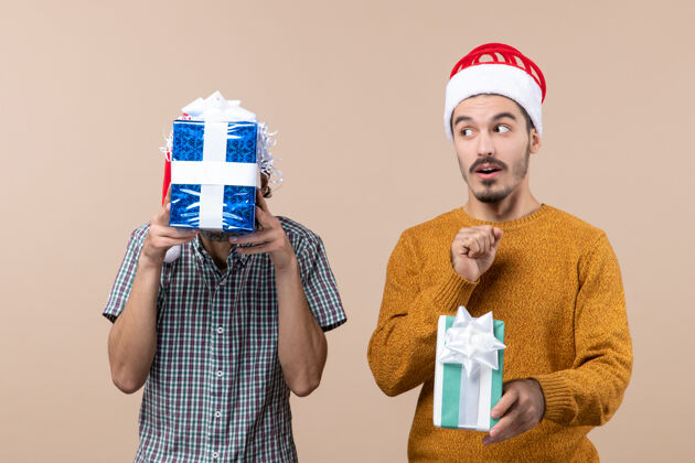 视图正面图两个戴着圣诞帽的惊讶的家伙手里拿着圣诞礼物 一个用礼物遮住脸 背景是米色的两个手持两个惊喜的家伙们