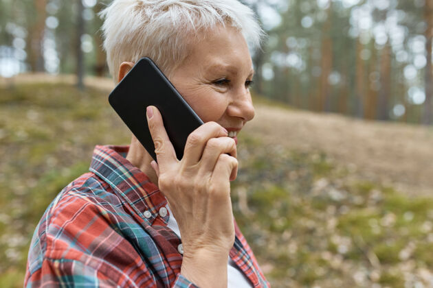 游客中年女性的肖像 手腕 穿着格子衬衫 在户外摆姿势 耳边拿着智能手机 谈吐愉快 喜欢在森林里散步徒步旅行退休风景