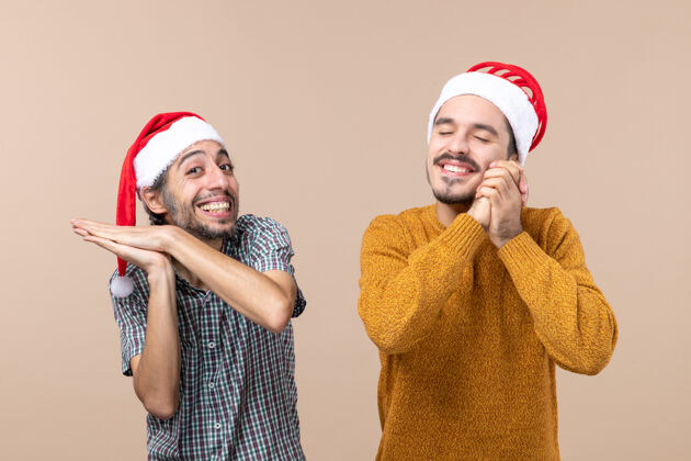 视图正面图两个戴着圣诞帽的快乐的家伙一个在拍手另一个闭着眼睛站在米色的孤立背景上许愿伙计们站着帽子