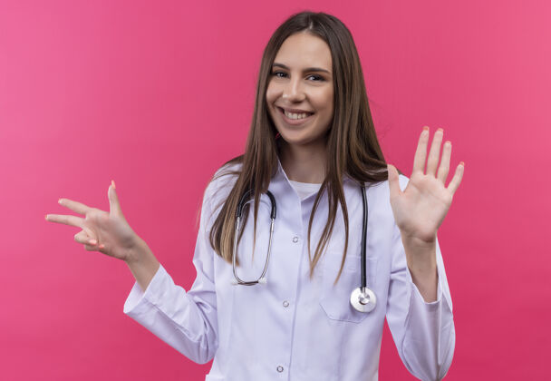 微笑微笑的年轻医生女孩穿着听诊器医用长袍 在孤立的粉红色背景上显示不同的数字展示医疗礼服