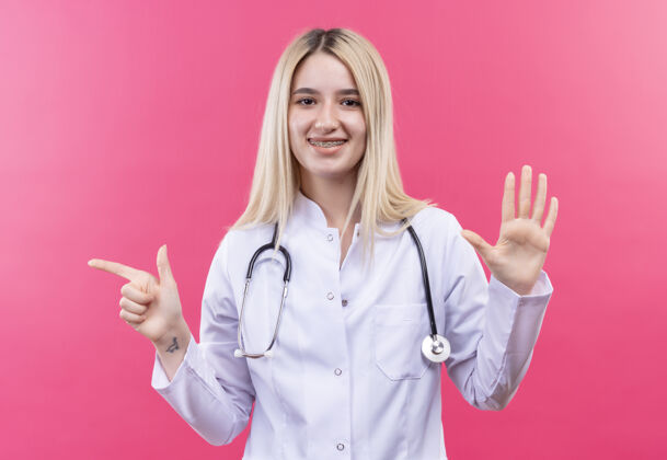 牙科微笑的医生年轻的金发女孩穿着医用长袍戴着听诊器和牙套 在孤立的粉色背景上显示出不同的手势听诊器穿着长袍