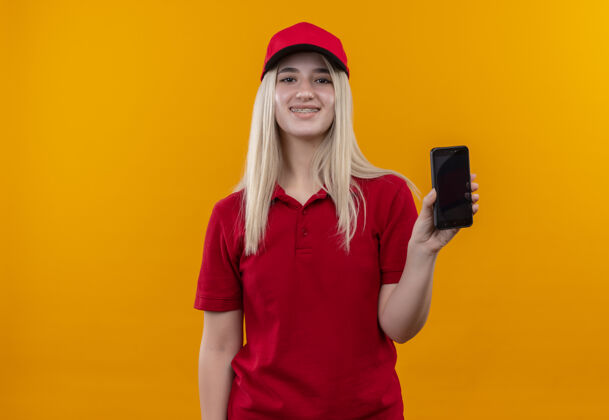 红色微笑的小女孩穿着红色t恤 戴着帽子 戴着牙套 在孤立的橙色背景下对着摄像机展示手机年轻帽子橙色