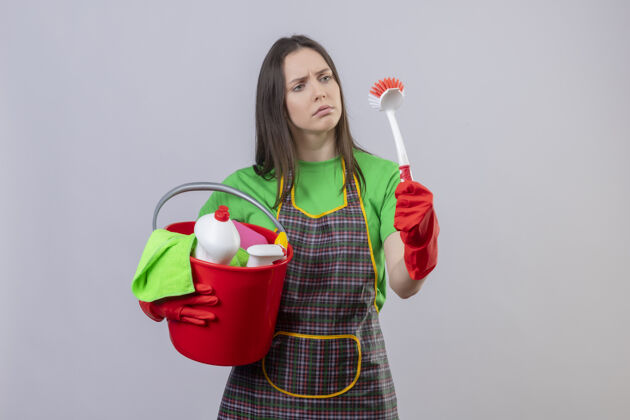 年轻悲伤的清洁年轻女孩穿着制服 戴着红色手套 手持清洁工具 在孤立的白色背景上看着手上的清洁刷工具悲伤手