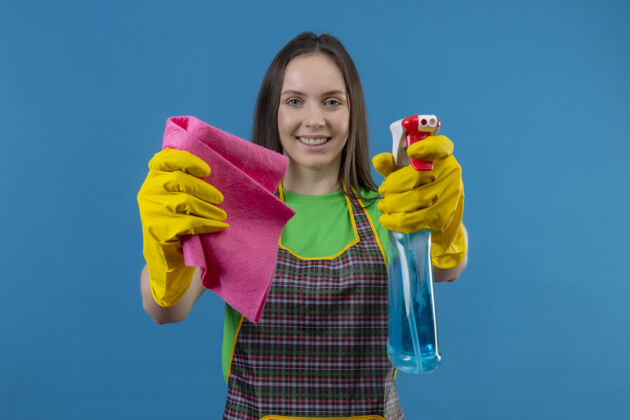 穿着微笑着清洗的年轻女孩穿着制服 戴着手套 拿着清洁喷雾和抹布对着隔离的蓝色背景上的摄像机年轻蓝色女孩