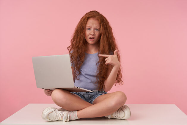 表情漂亮的少女 迷人的卷发 手举食指 交叉着双腿坐在粉色背景下 手里拿着一台现代化的笔记本电脑孩子腿肖像