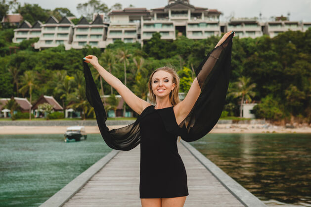 风穿着黑色连衣裙的美丽迷人的女人在豪华度假酒店的码头上摆姿势 暑假 热带海滩天堂奢华优雅