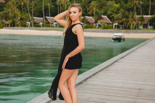 赤脚穿着黑色连衣裙的美丽迷人的女人在豪华度假酒店的码头上摆姿势 暑假 热带海滩天堂度假胜地长袍