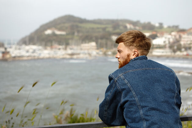 站立帅气的留着胡子的年轻人穿着时髦的蓝色的后画像休闲海滩海岸