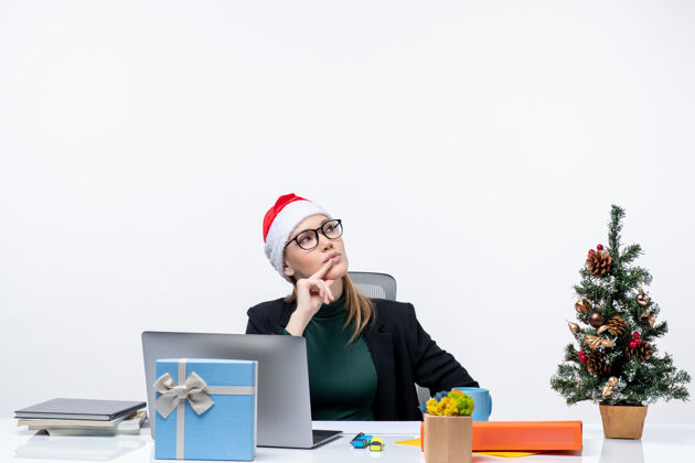 圣诞老人一位惊讶的商务女士戴着圣诞老人帽坐在一张桌子旁 桌子上放着圣诞树和礼物 在白色背景上指着左边桌子惊讶办公室
