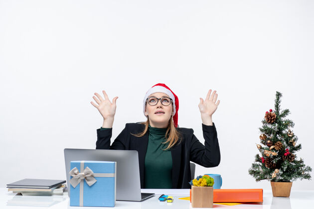 笔记本电脑一位戴着圣诞老人帽子的商务女士坐在一张桌子旁 桌子上有一棵圣诞树 上面有一件礼物 背景是白色的帽子商务女士笔记本电脑