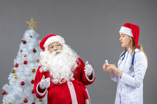 圣诞老人圣诞老人和女医生的前视图 女医生在灰色的墙上拿着测温仪节日测量女性