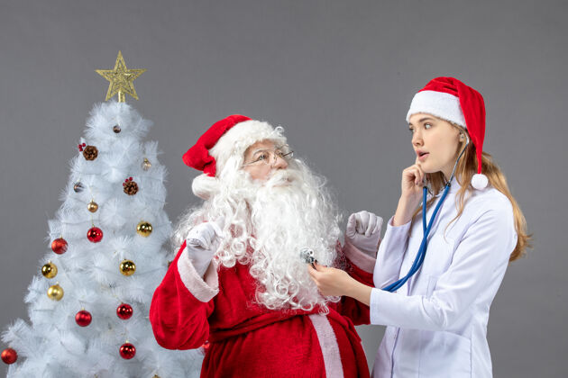 健康圣诞老人和女医生的前视图 女医生正在灰色墙上检查他的健康状况快乐圣诞老人正面