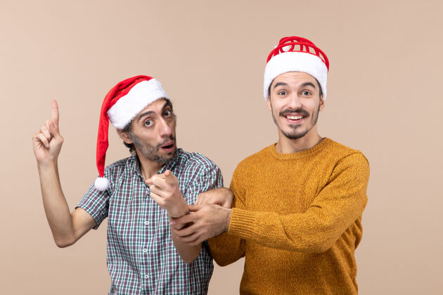 展示正面图两个带着圣诞帽的微笑的家伙一个在展示什么 另一个在米色孤立背景上牵着他的手手拿着前面
