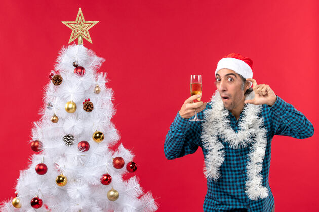 新的圣诞心情与情绪化的年轻人 戴着圣诞老人帽 穿着蓝色条纹衬衫 手拿一杯葡萄酒 在圣诞树旁仔细聆听庆祝装饰举行