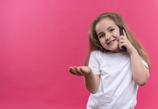 小微笑着的小女生穿着白色t恤在电话里讲话 在孤立的粉色背景下伸出手向前走向前微笑女孩