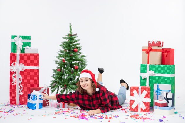 周围正面图：年轻的女性躺在圣诞礼物和小圣诞树周围 背景是白色的寒冷的女人圣诞节新年下雪树冬青圣诞