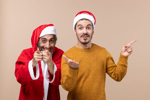 手指正面图两个戴着圣诞帽的朋友都用手指在米色的孤立背景上展示相机米色圣诞老人肖像