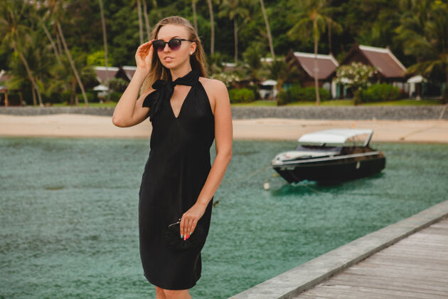 魅力奢华性感迷人的女人穿着黑色礼服在豪华度假酒店的码头上摆造型 戴着墨镜 暑假 热带海滩海滩天堂摆姿势