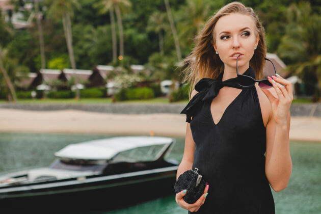 夏天奢华性感迷人的女人穿着黑色礼服在豪华度假酒店的码头上摆造型 戴着墨镜 暑假 热带海滩热带游艇异国情调