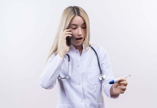 体温计惊讶的医生穿着医用长袍戴着听诊器的年轻金发女孩在电话里说话看着她手上孤立的白色背景上的温度计看说话长袍