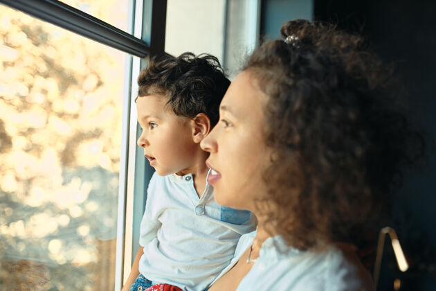 幼儿快乐的年轻拉丁母亲呆在家里的室内肖像站在窗边 抱着学龄前的儿子 透过玻璃向外看小好奇小