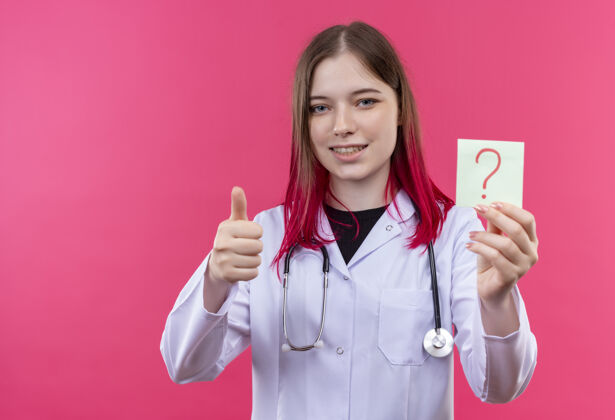 拇指微笑的年轻医生女孩穿着听诊器医用长袍拿着纸问号她的拇指上粉红色的孤立背景纸听诊器问题