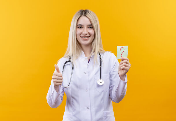 微笑微笑的医生年轻的女孩穿着医用长袍戴着听诊器戴着牙套拿着纸问号在孤立的黄色背景上竖起大拇指牙科支架黄色