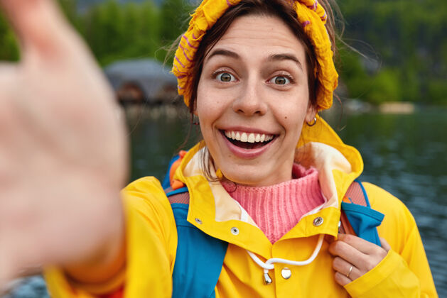 高兴微笑的女人伸手自拍的特写镜头戴着黄色头带和雨衣呼吸着新鲜空气 站在河边风景湖泊反应