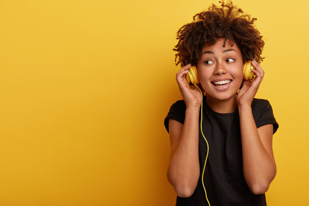 卷发快乐的黑皮肤女人戴着有线耳机 享受着美妙的音乐 目光移开 露出牙齿般的微笑耳机好音频