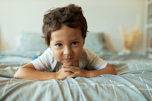 毯子健康迷人的3岁拉丁幼儿躺在皱巴巴的床单上双手紧握在他面前 有着好奇有趣的表情脸乐趣睡眠