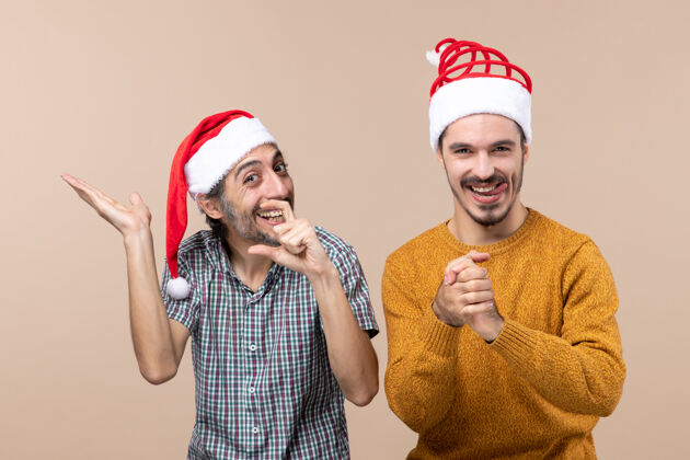 展示正面图：两个戴着圣诞帽的笑脸 一个在展示什么 另一个在米色孤立的背景上双手合十男帽子看
