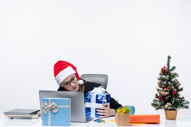 圣诞树带着圣诞心情的女商人戴着圣诞老人的帽子 戴着眼镜坐在一张桌子旁 把礼物和圣诞树装饰在它的白色背景上人商务圣诞